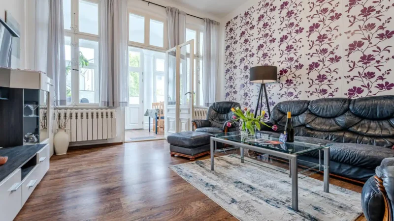 Auf diesem Bild sehen Sie das Wohnzimmer mit Anbauwand, Fernseher, Couch-Garnitur und Blick auf die geschlossene Veranda mit großer Fensterfront in der Ferienwohnung 75 Quadratmeter im Erdgeschoss des Ferienhauses der Villa See-Eck im Seebad Ahlbeck auf der Insel Usedom von https://aufnachusedom.de.