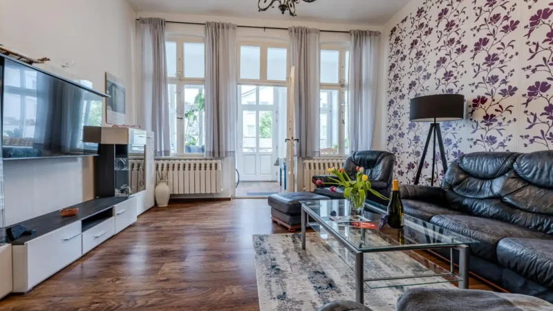 Auf diesem Bild sehen Sie das Wohnzimmer mit Anbauwand, Fernseher, Couch-Garnitur und Blick auf die geschlossene Veranda mit großer Fensterfront in der Ferienwohnung 75 Quadratmeter im Erdgeschoss des Ferienhauses der Villa See-Eck im Seebad Ahlbeck auf der Insel Usedom von https://aufnachusedom.de - Bild 2.